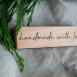 A rustic wooden sign with 'Handmade with Love' text on linen, accented by green foliage.